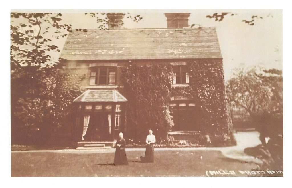 Historic photograph of Armitage Cottage, Rugeley Road, showing the ivy-covered two-storey house, glazed entrance porch, gardens and two women standing on the lawn.