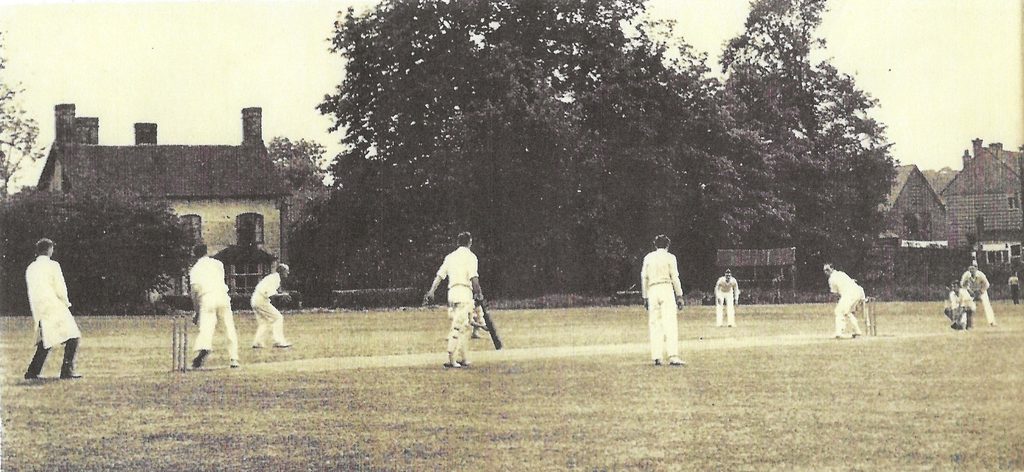 Historic photograph of a cricket match being played on the former Armitage cricket ground, with players in white clothing and houses and mature trees in the background.